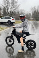 Motocyklista w kasku ochronnym siedzący na czarnym elektrycznym motocyklu Tuttio Soleil 01 SE na mokrej, asfaltowej drodze osiedlowej podczas opadów deszczu lub śniegu.