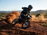 Dynamiczne ujęcie motocyklisty w pełnym stroju ochronnym pokonującego ostry zakręt na piaszczystym torze na czarnym elektrycznym motocyklu crossowym Tuttio Soleil 01 SE, wzbijając tumany kurzu w słoneczny dzień.