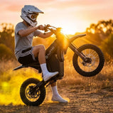 Motocyklista w kasku wykonujący jazdę na tylnym kole (wheelie) na czarnym elektrycznym motocyklu Tuttio Soleil 01 SE na piaszczystej drodze o zachodzie słońca.